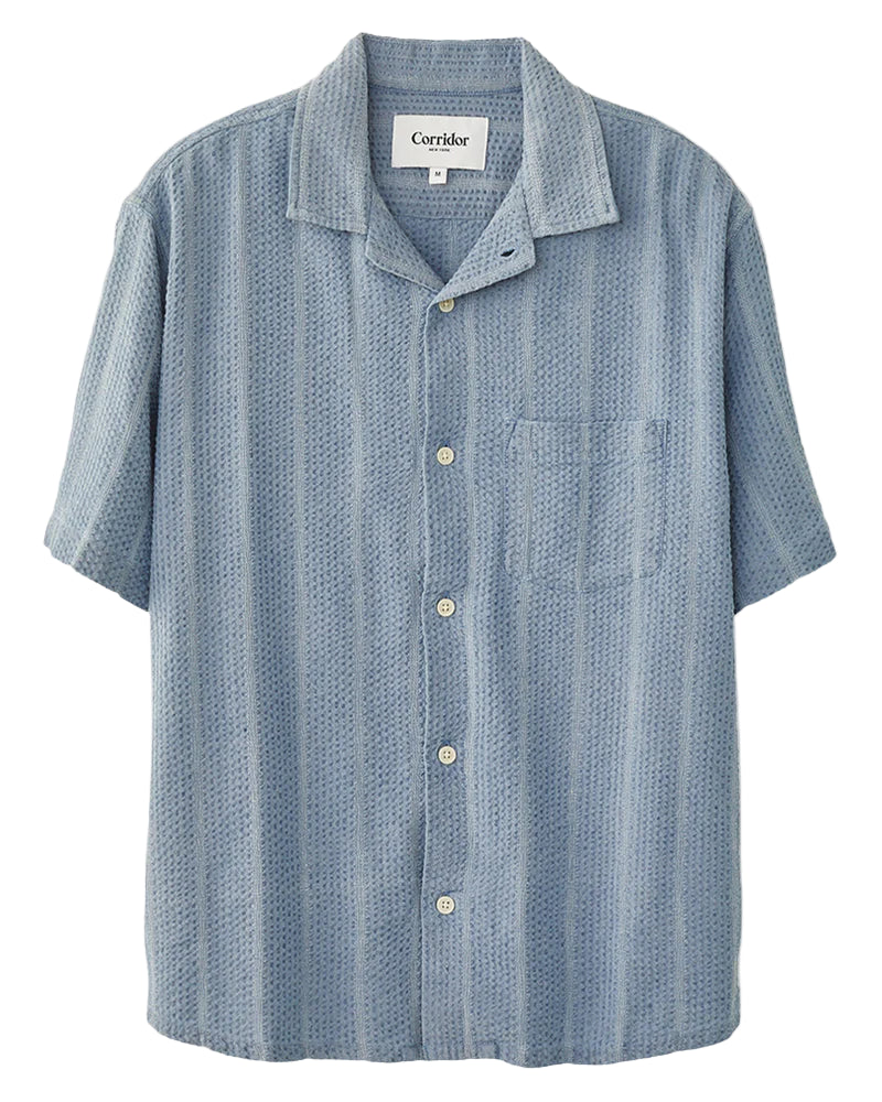 A striped seersucker short-sleeved shirt in blue and white, with a camp collar and corozo buttons, displayed on a white background.