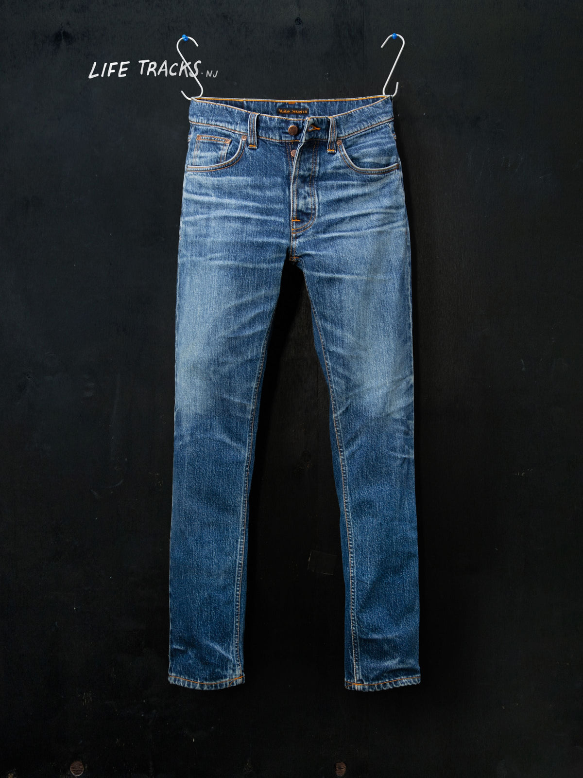 Nudie Jeans Solid Ollie Life Tracks in a mid-blue wash, hung against a dark background with 'Life Tracks' written above.