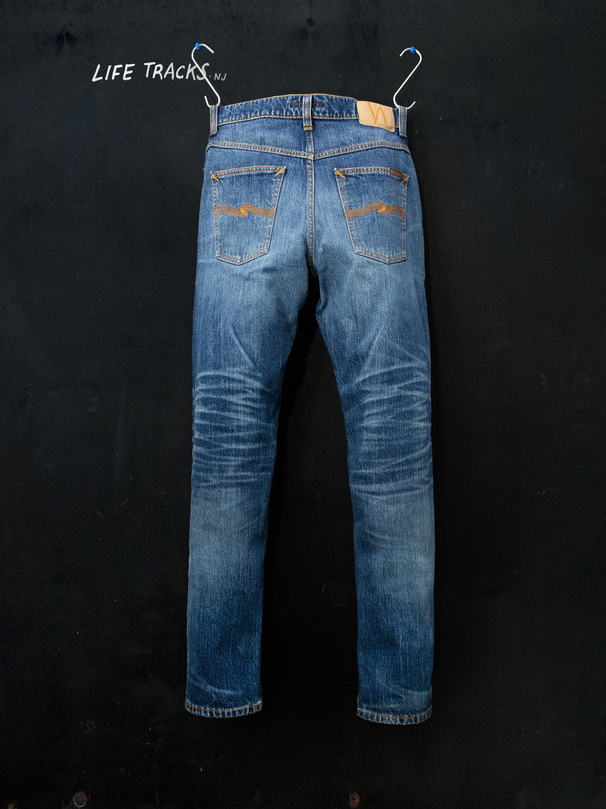 Nudie Jeans Solid Ollie Life Tracks, mid-blue wash, back view hanging on a black wall with 'Life Tracks' text.