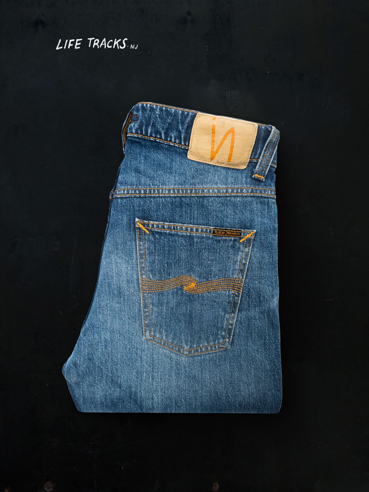 Nudie Jeans Solid Ollie Life Tracks in a mid-blue wash, folded and laid flat on a black background with 'Life Tracks' text.