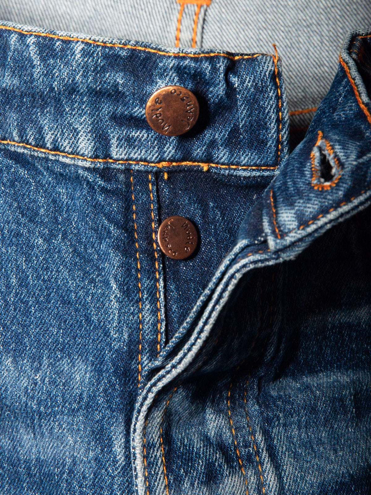 Nudie Jeans Solid Ollie Life Tracks, a close-up of the copper button fly and orange stitching on the faded blue denim.